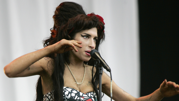 A white woman wearing a gold necklace and geometric triangle dress holds her arms out while singing into a microphone.