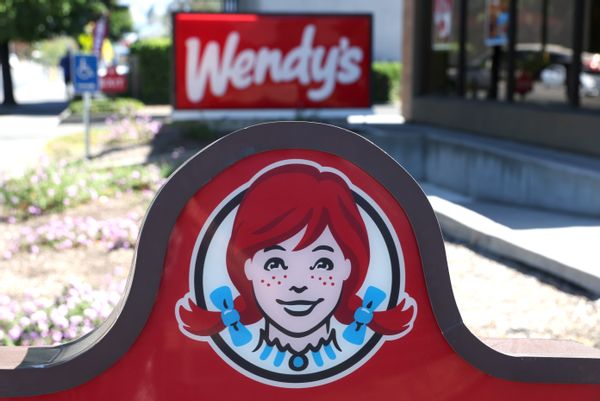 A white girl with red hair  in pigtails smiles on a red sign. Behind it is another sign that says Wendy's. 